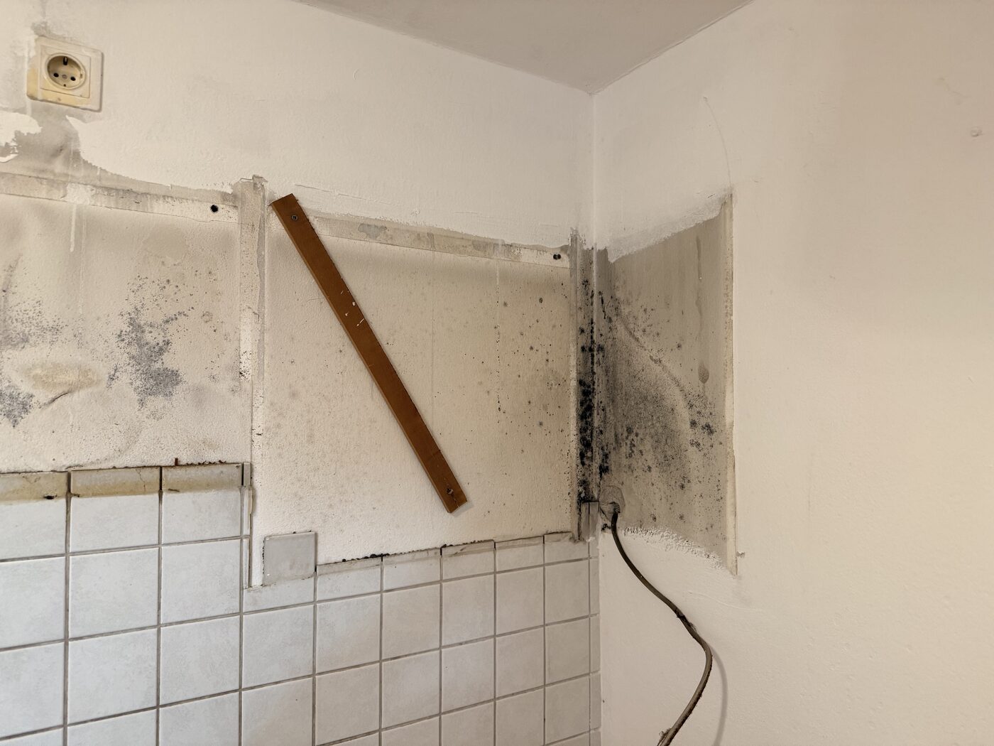 Wandecke mit dunklen Feuchteschäden über einem alten Fliesenspiegel