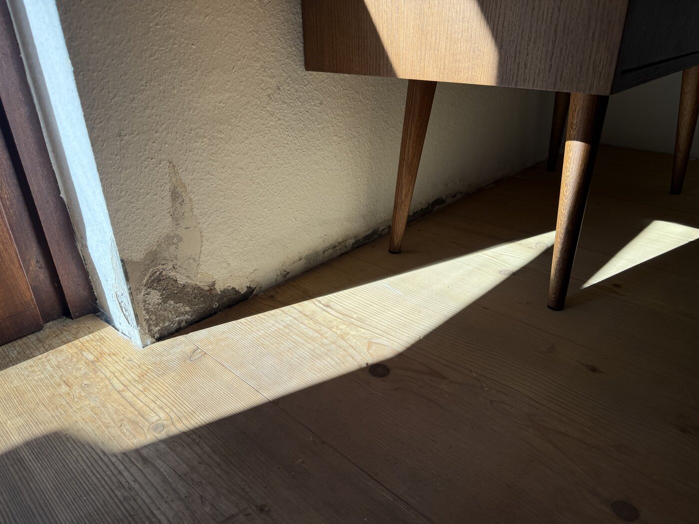 Dunkle Feuchtespuren im Sockelbereich einer Wand, hinter einem Möbelstück